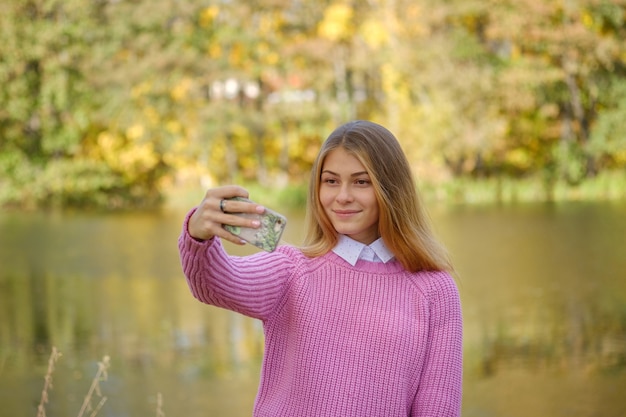 Junges europäisches Mädchen nimmt ein Selfie auf einem Smartphone über einer schönen Herbstlandschaft auf dem See