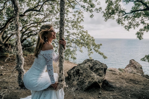 Junges europäisches schönes Mädchen in weißem Brautkleid, das auf einer Schaukel in Waldbäumen am Meeresstrandhintergrund posiert Exotische Hochzeitsfotografie-Ideen