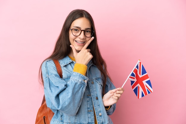 Junges französisches Mädchen, das eine britische Flagge lokalisiert auf rosa Hintergrund glücklich und lächelnd hält