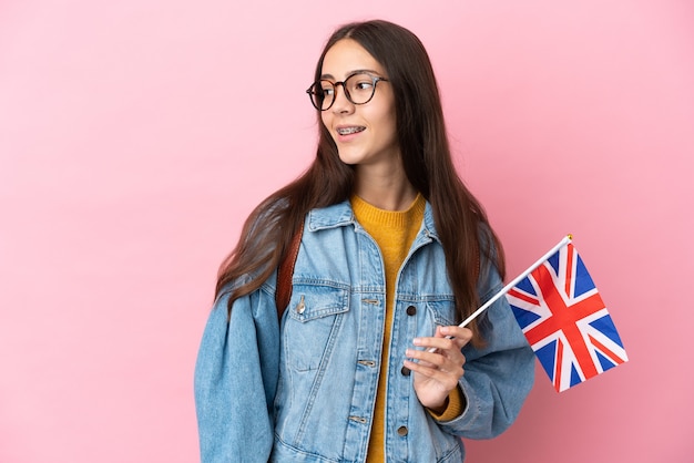 Junges französisches Mädchen, das eine britische Flagge lokalisiert auf rosa Hintergrund hält, der zur Seite schaut und lächelt