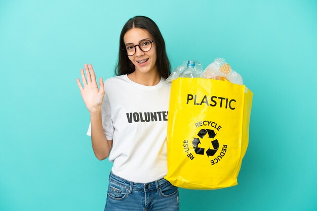 Junges französisches Mädchen, das eine Tasche voller Plastikflaschen hält, um den Gruß mit der Hand mit glücklichem Ausdruck zu recyceln