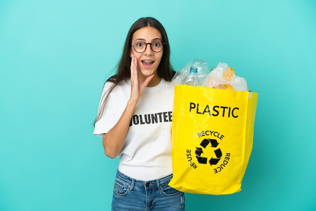 Junges französisches Mädchen, das eine Tasche voller Plastikflaschen hält, um sie mit Überraschung und schockiertem Gesichtsausdruck zu recyceln