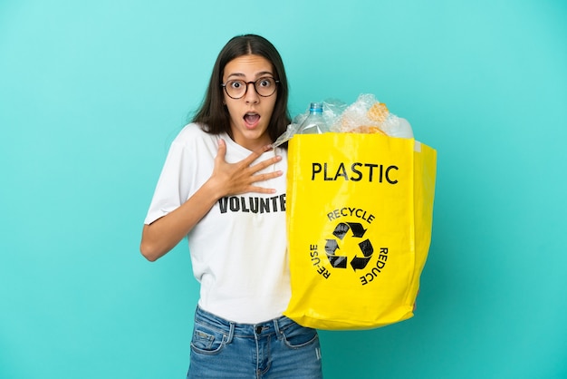 Junges französisches Mädchen, das eine Tasche voller Plastikflaschen hält, um überrascht und schockiert zu recyceln, während es richtig schaut