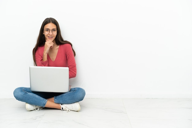 Junges französisches Mädchen, das mit ihrem Laptop mit Brille auf dem Boden sitzt und lächelt