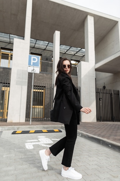 Junges Frauenmode-Modell in schwarzer Vintage-Kleidung mit Handtasche in modischer Sonnenbrille geht in der Nähe eines modernen Gebäudes in der Stadt. Cooles amerikanisches Mädchen im Freizeitkleid, das in der Straße aufwirft. Stilvolle Damenbekleidung.