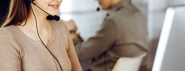 Junges freundliches Mädchen in Headsets spricht mit einem Firmenkunden, während sie zusammen mit ihrer Kollegin am Schreibtisch in einem modernen Büro sitzt. Call-Center-Betreiber bei der Arbeit.