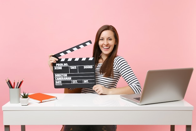 Junges fröhliches Mädchen, das klassischen schwarzen Film hält, der eine Klappe macht und am Projekt arbeitet, während er mit Laptop im Büro sitzt