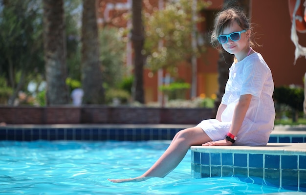 Junges fröhliches Mädchen, das sich an sonnigen Sommertagen auf der Seite des Swimmingpools mit klarem blauem Wasser ausruht Tropisches Urlaubskonzept