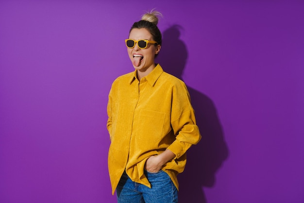 Junges fröhliches Mädchen mit gelbem Hemd und Sonnenbrille vor violettem Hintergrund