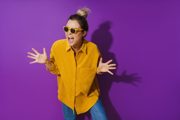 Junges fröhliches Mädchen mit gelbem Hemd und Sonnenbrille vor violettem Hintergrund