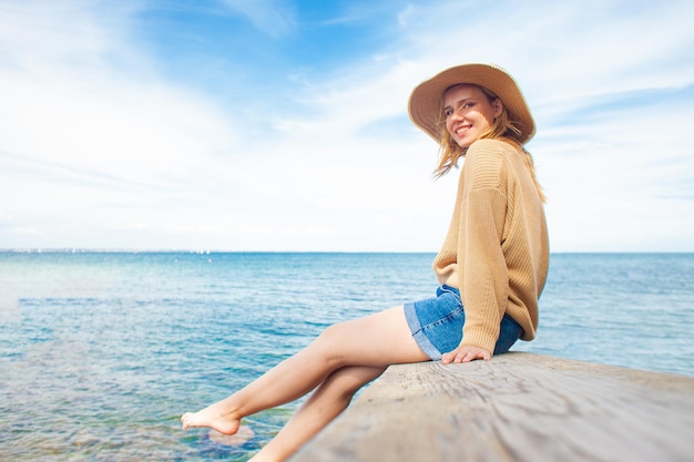 Junges fröhliches Mädchen sitzt barfuß auf dem Pier und lächelt auf dem Hintergrund des Meeres
