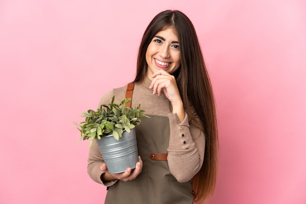 Junges Gärtnermädchen, das eine Pflanze isoliert auf rosa Hintergrund hält und zur Seite schaut und lächelt