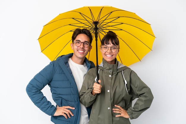 Junges gemischtes Paar hält einen Regenschirm isoliert auf weißem Hintergrund posiert mit den Armen an der Hüfte und lächelt