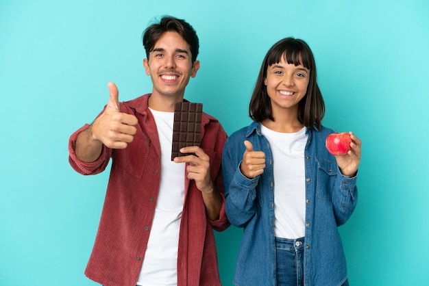 Junges gemischtes Rassenpaar, das Apfel und Schokolade isoliert auf blauem Hintergrund hält und eine Daumen hoch Geste gibt, weil etwas Gutes passiert ist