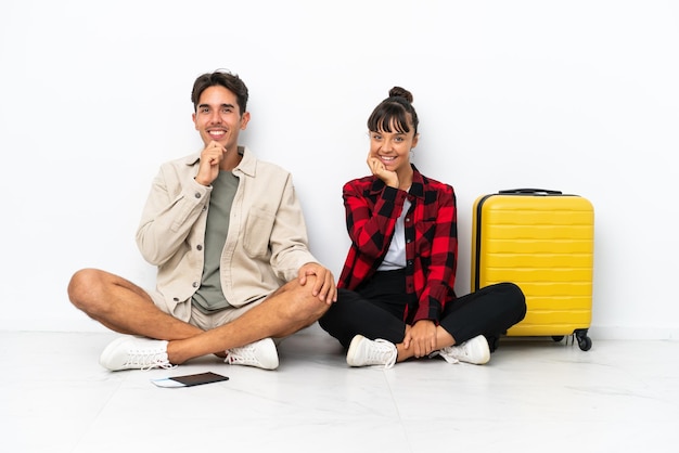 Junges gemischtes Reisendespaar, das auf dem Boden lokalisiert auf weißem Hintergrund sitzt und mit einem süßen Ausdruck lächelt