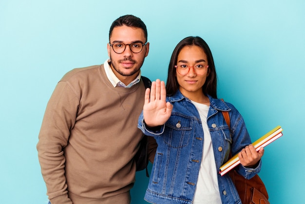 Junges gemischtes Studentenpaar isoliert auf blauem Hintergrund, das mit ausgestreckter Hand steht und Stoppschild zeigt und Sie verhindert.