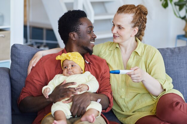 Junges gemischtrassiges Paar, das auf Sofa mit Baby sitzt und Schwangerschaftstest macht