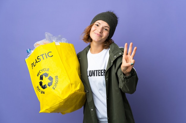 Junges georgianisches Mädchen, das eine Tasche voller Plastikflaschen hält, um glücklich zu recyceln und drei mit den Fingern zu zählen