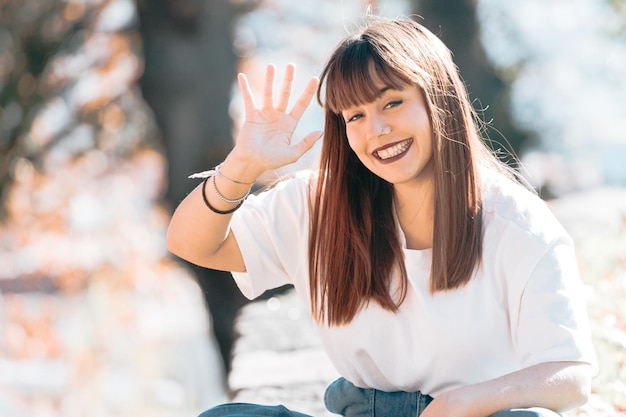 Junges glückliches aktives Mädchen, das beim Sitzen zur Kamera grüßt und ein großes Zähnelächeln lächelt. Platz kopieren. Weißes Hemd und trendiges modernes Outfit. Glückliches Lächeln Zahnarztkonzept, Mundgesundheit. Körper positiver Geist