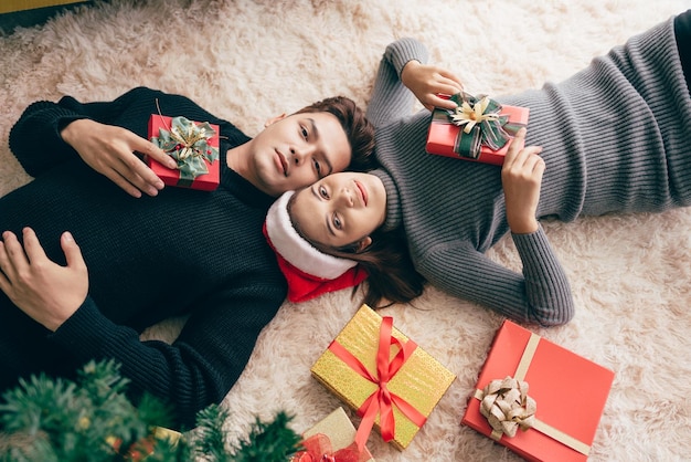 Junges glückliches asiatisches Paar, das einen Pullover trägt, der sich zusammen hinlegt und während der Weihnachtsferien Geschenke mit vielen Geschenkboxen auf dem Teppich im Wohnzimmer hält