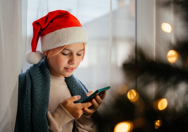 Junges glückliches jugendlich Mädchen in rotem Sankt-Hut mit Handy mit Weihnachtsbaum im Vordergrund