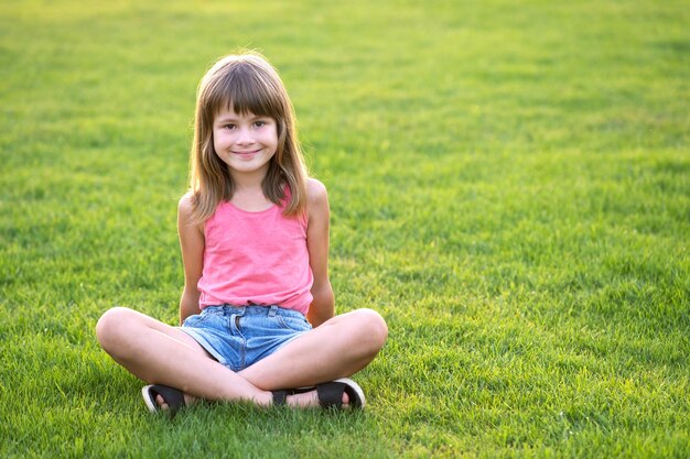 Junges glückliches Kindermädchen, das beim Sitzen auf grünem Grasrasen an einem warmen Sommertag stillsteht.