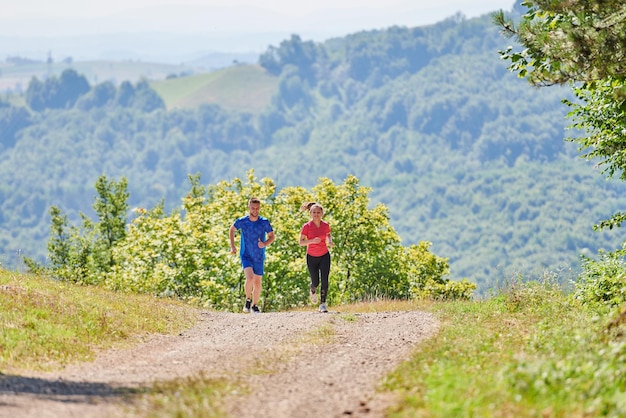 junges glückliches paar, das einen gesunden lebensstil genießt, während es auf einer landstraße durch den schönen sonnigen wald joggt, übungs- und fitnesskonzept