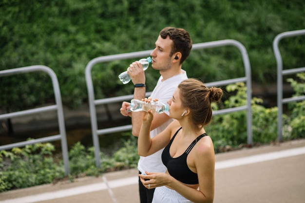 Junges glückliches Paar, das im Stadtpark mit einer Flasche Wasser in den Händen läuft, gemeinsamer Sport, Fröhlichkeit, Stadtsport gesunder Lebensstil, gemeinsame Fitness am Sommerabend, Läufer, Trinkwasser, Durst