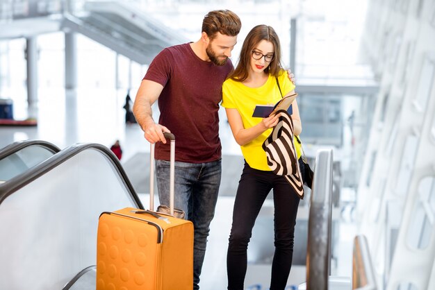 Junges glückliches Paar, das während ihres Sommerurlaubs mit Gepäck auf der Rolltreppe zum Abflugbereich des Flughafens aufsteht