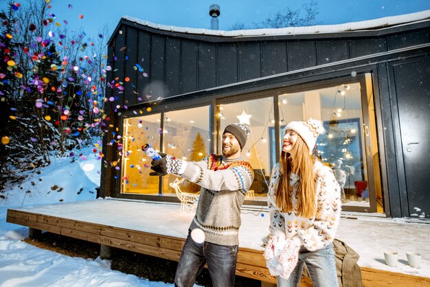 Junges glückliches Paar in Pullovern, die Winterferien vor einem wunderschön dekorierten Haus in den Bergen feiern
