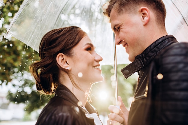 Junges glückliches Paar küssen unter einem transparenten Regenschirm auf einer Stadtstraße. Nahaufnahmeporträt von Mann und Frau. Liebespaar, das unter einem Regenschirm lächelt. Sonniger Herbsttag in der Stadt.