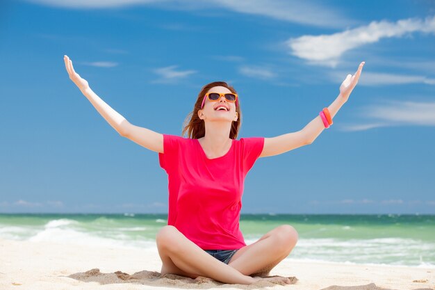 Junges glückliches rothaariges Mädchen in der Sonnenbrille mit den Händen oben auf Sommerzeitküste des Meeres.