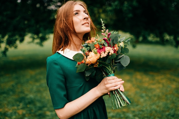 Junges glückliches rothaariges Mädchen mit Blumenstrauß der Sommerblumen, die inneren Frieden im grünen Kleid an der Natur genießen. Hübsches Frauenporträt im Freien. Schönes lächelndes Gesicht.