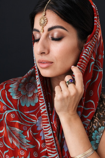 Junges hinduistisches Mädchen, das traditionelles indisches Saree-Kleid und ethnischen Schmuck trägt, der ihren Kopf mit Dupatta-Schal bedeckt, isoliert über schwarzer Wand?