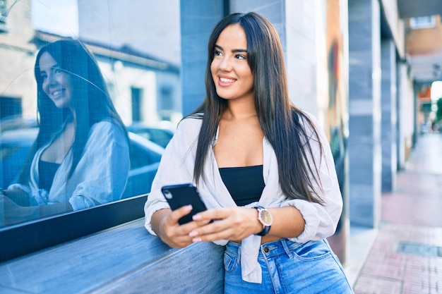 Junges hispanisches Mädchen lächelt glücklich mit Smartphone in der Stadt