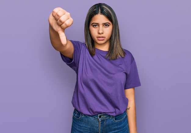 Junges hispanisches Mädchen trägt lässiges lila T-Shirt und sieht unglücklich und wütend aus, zeigt Ablehnung und Negativität mit der Daumen-nach-unten-Geste. schlechter Ausdruck.