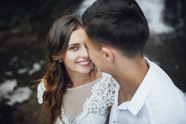 Junges Hochzeitspaar des hübschen Mädchens oder der Frau mit den Haaren im weißen Brautkleid und im hübschen Mann nahe dem schönen großen Wasserfall