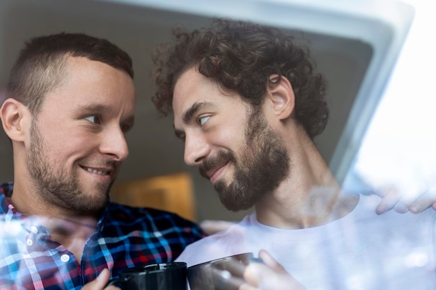 Junges homosexuelles paar in der liebe, die aus dem fenster schaut zwei junge androgyne männer, die zusammen lächeln und kaffee trinken