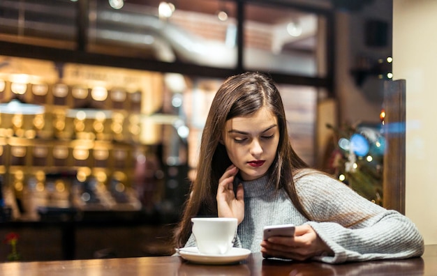 Junges hübsches brünettes Mädchen trägt grauen Pullover und surft per Smartphone im Internet, das in der Nähe des Caféfensters sitzt