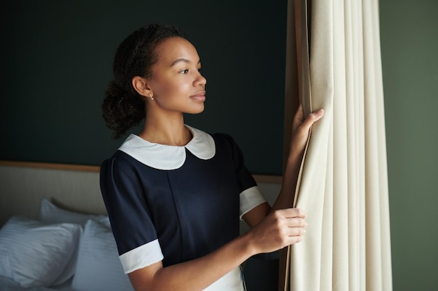Junges hübsches Hausmädchen im dunkelblauen Kleid, das Vorhänge im Hotelzimmer öffnet