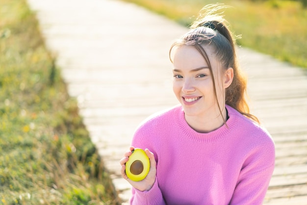 Junges hübsches Mädchen, das draußen eine Avocado hält und viel lächelt