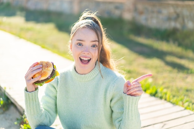 Junges hübsches Mädchen, das draußen einen Burger hält, überrascht und zeigt mit dem Finger zur Seite