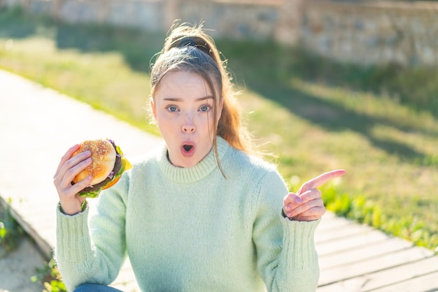 Junges hübsches Mädchen, das draußen einen Burger hält, überrascht und zeigt Seite