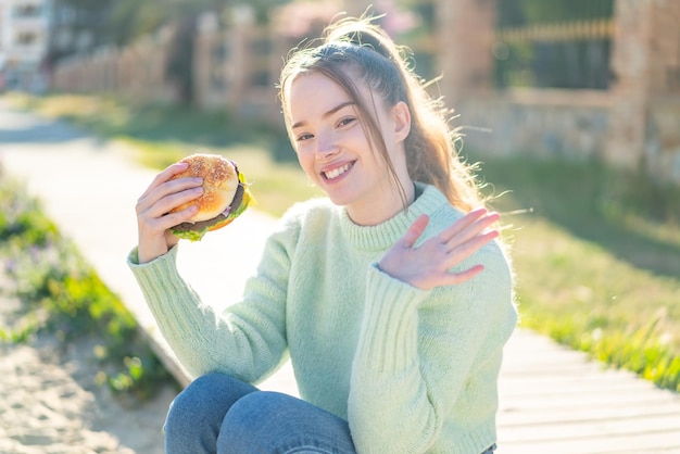 Junges hübsches Mädchen, das draußen einen Burger hält und mit der Hand mit glücklichem Ausdruck salutiert