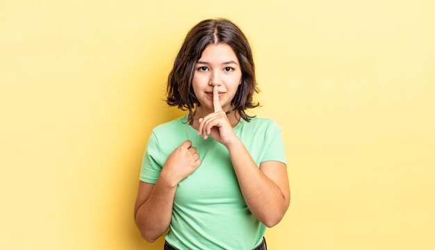 Junges hübsches Mädchen, das ernst aussieht und mit dem Finger auf die Lippen gedrückt wird, um Stille oder Ruhe zu fordern, ein Geheimnis zu bewahren