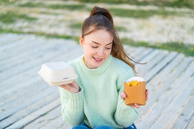 Junges hübsches Mädchen im Freien, das gebratene Pommes und Takeaway-Box-Essen und Denken nimmt