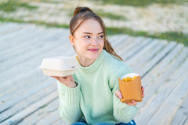 Junges hübsches Mädchen im Freien, das gebratene Pommes und Takeaway-Box-Essen und Denken nimmt