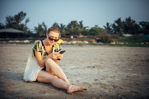 Junges hübsches Mädchen im kurzen Kleid und in der Sonnenbrille, die auf dem Sand sitzen und Smartphone halten. Frau und glückliche Zeit im Urlaub