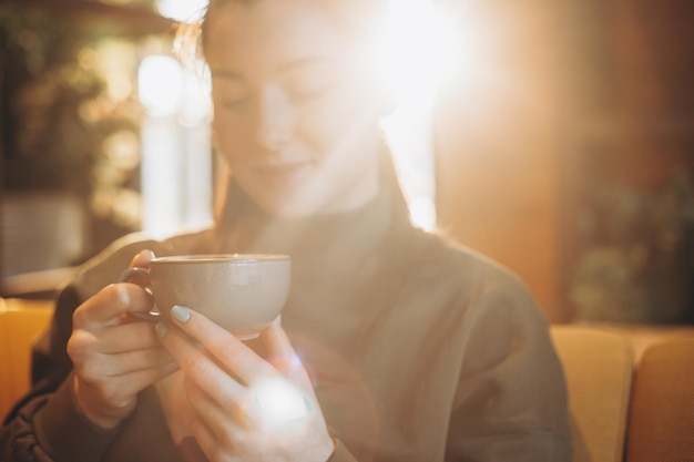 Junges hübsches Mädchen mit einer großen Tasse süßen Kaffees sitzt auf einem Stuhl in ihrem Lieblingscafé und trinkt ihren täglichen Morgenkaffee und spricht mit einer Freundin