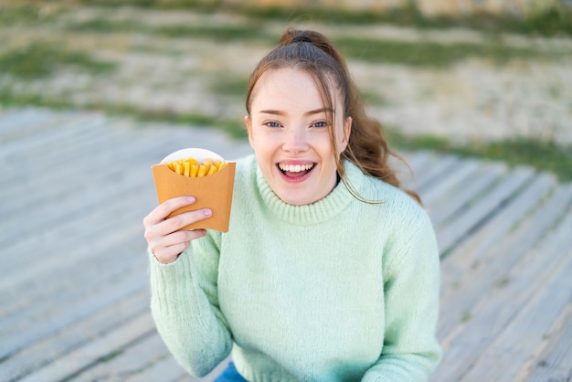 Junges hübsches Mädchen mit gebratenen Chips im Freien mit Überraschung und schockiertem Gesichtsausdruck